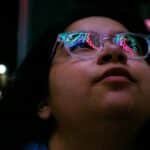 A young woman looks up reflecting colorful neon lights in her glasses at night.