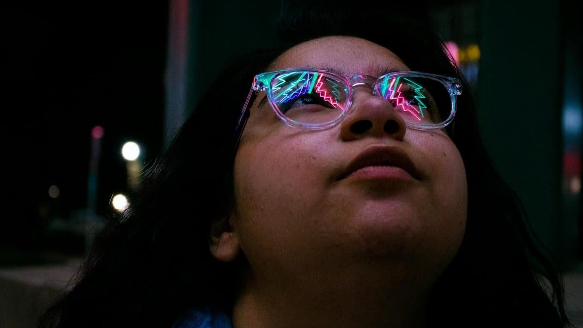 A young woman looks up reflecting colorful neon lights in her glasses at night.