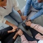 Four people sit in a circle holding hands, emphasizing unity and support in a therapy session.