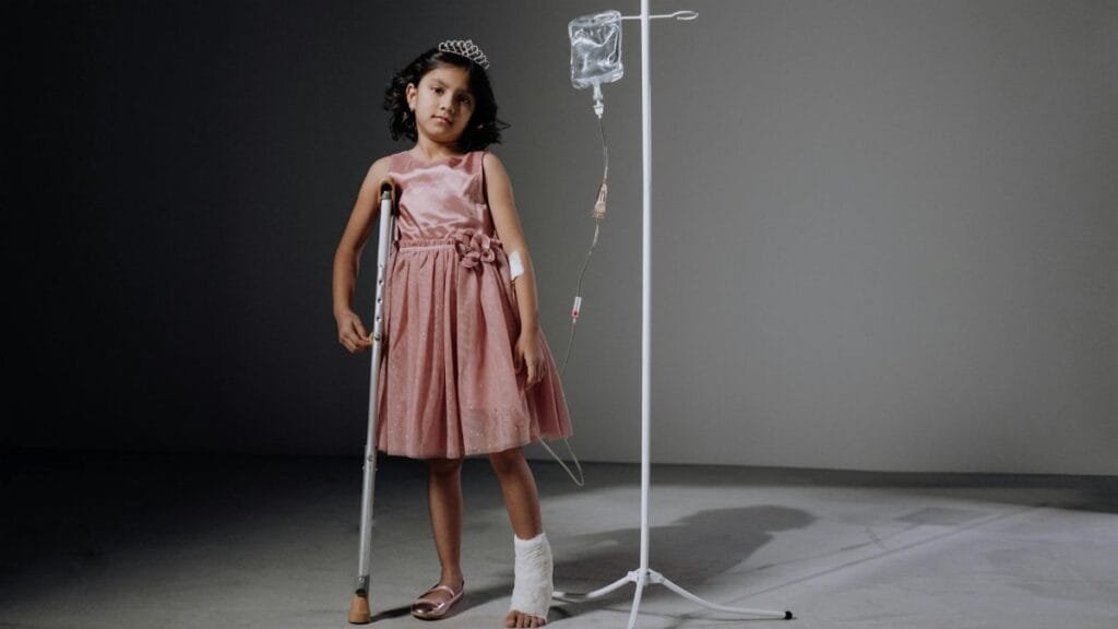 Injured girl in pink dress using crutches next to an IV stand in a studio setting.