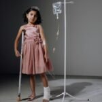 Injured girl in pink dress using crutches next to an IV stand in a studio setting.