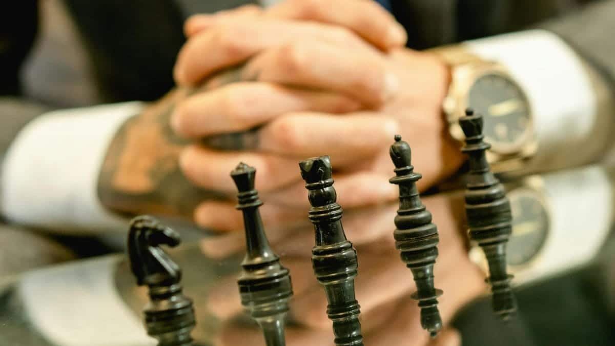 Close-up of chess pieces on a glass table with a person's clasped hands in the background.