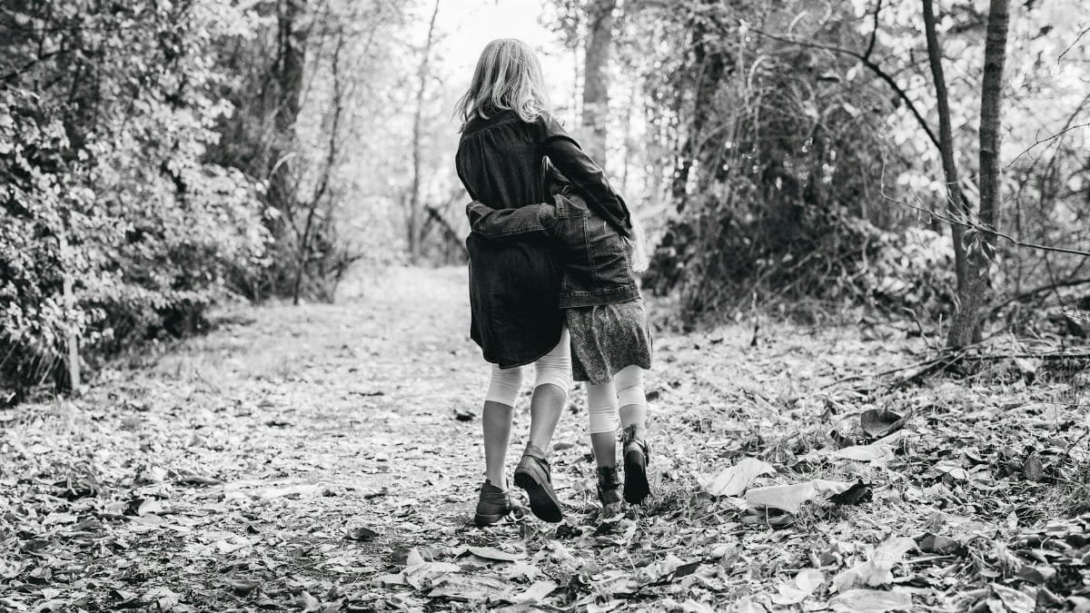 Black and white photo of sisters embracing on a forest walk surrounded by fallen leaves.