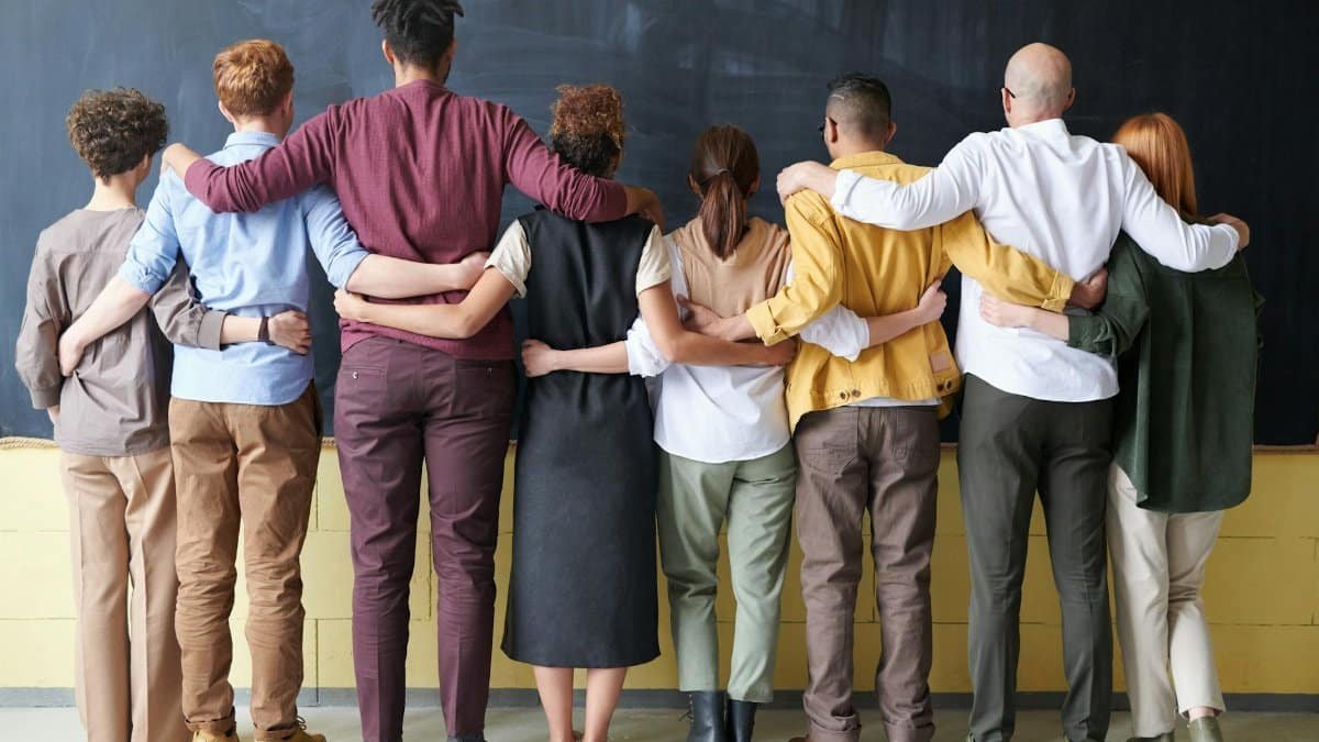 A diverse group of adults in casual outfits hugging in front of a chalkboard, symbolizing teamwork.