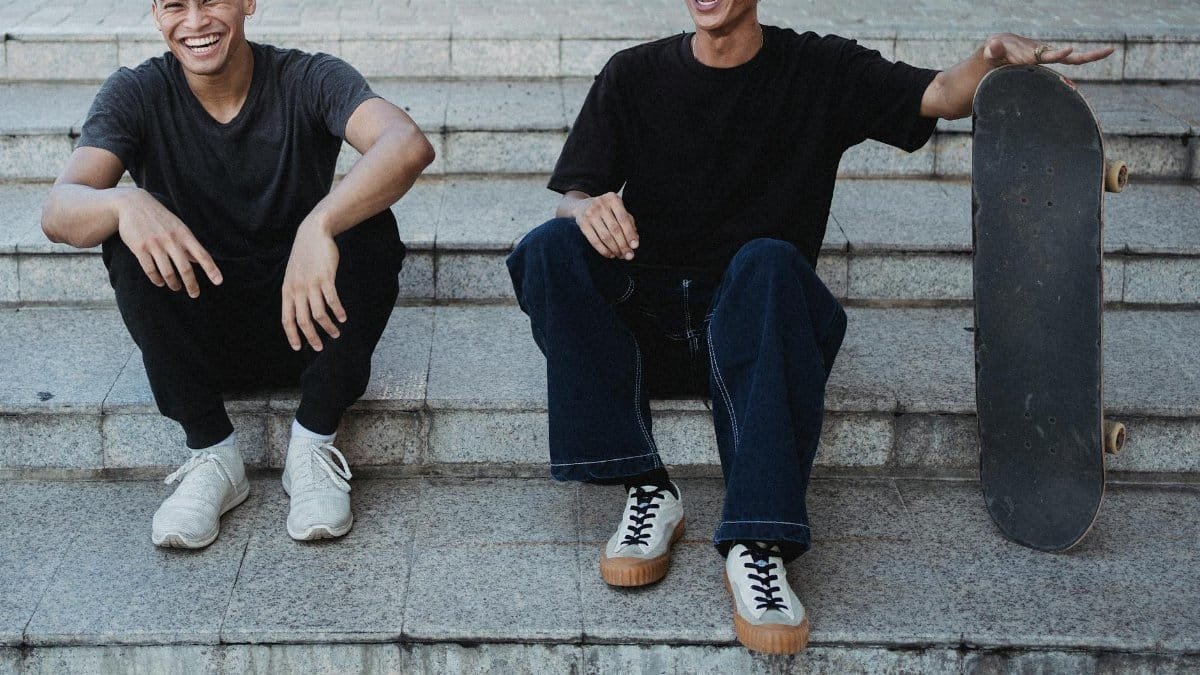 Two cheerful young men with a skateboard sitting on steps outdoors.