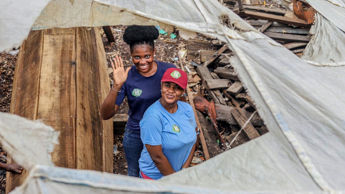 Two women smiling and waving, standing among construction debris, showing resilience.