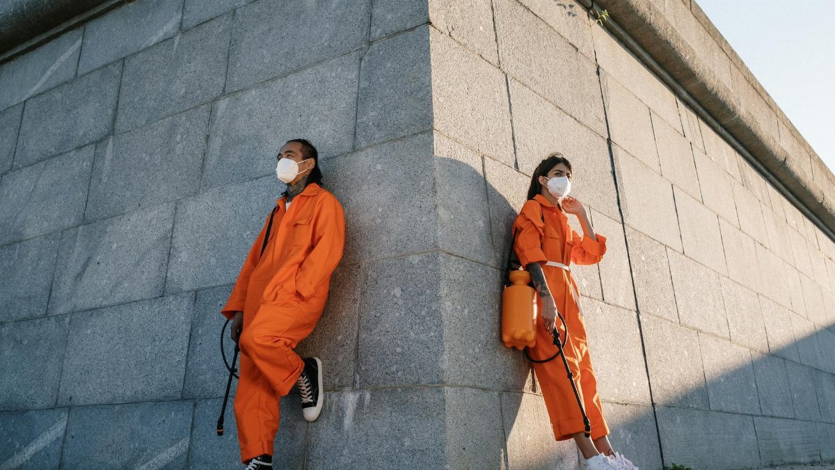 Two people in orange jumpsuits and masks leaning on a city wall.