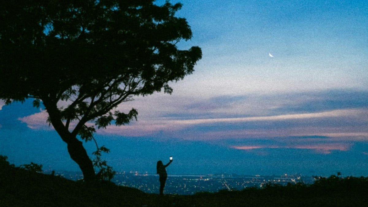 A silhouette of a person taking a photo against a cityscape during dusk with a visible crescent moon.