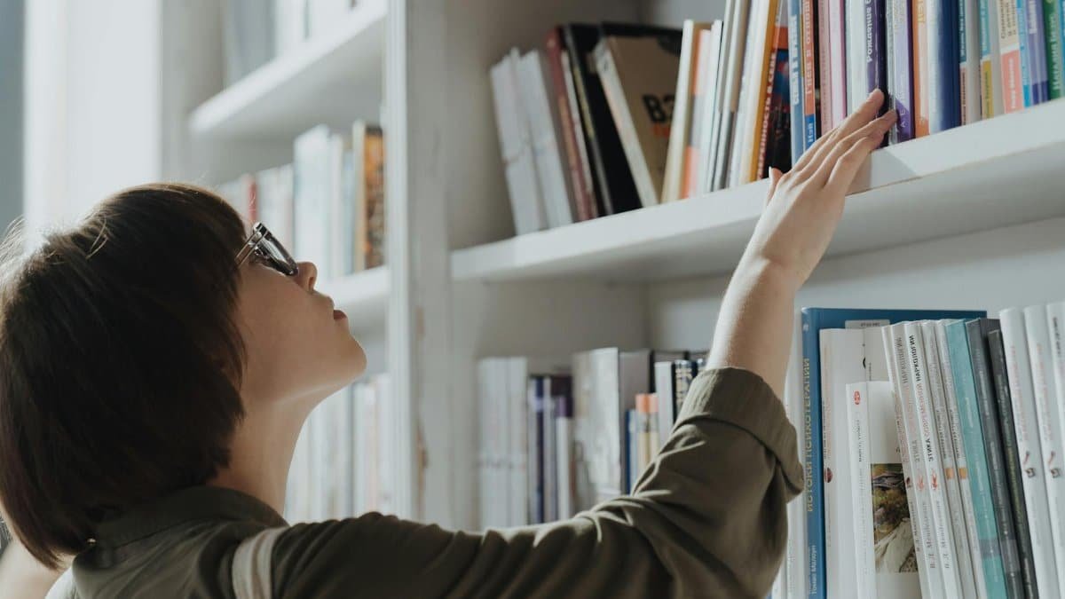 A woman wearing glasses browsing books on a library shelf, seeking knowledge and information.