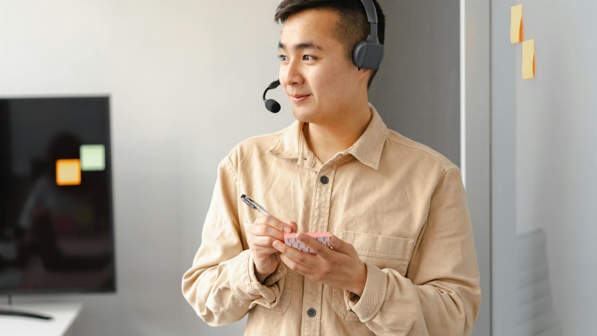 Smiling Asian man with headset holding notes, representing customer service in an office.