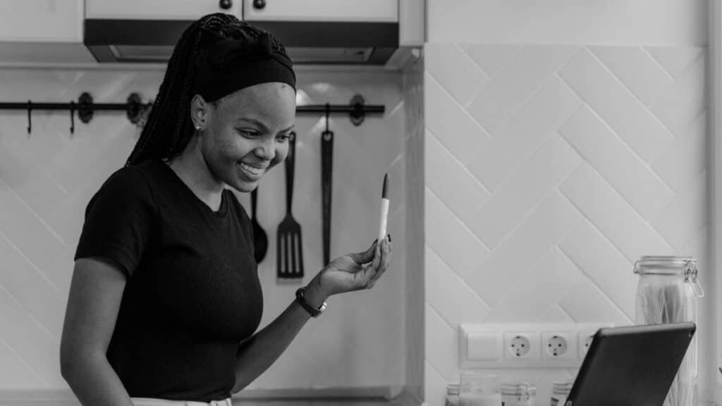 A joyful woman smiling at a positive pregnancy test during a video call in a modern kitchen.