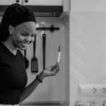 A joyful woman smiling at a positive pregnancy test during a video call in a modern kitchen.