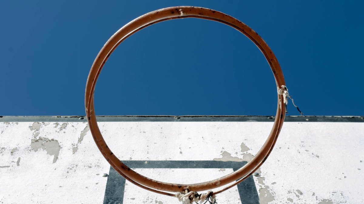 An old, rusted basketball hoop on a weathered backboard under a clear blue sky.
