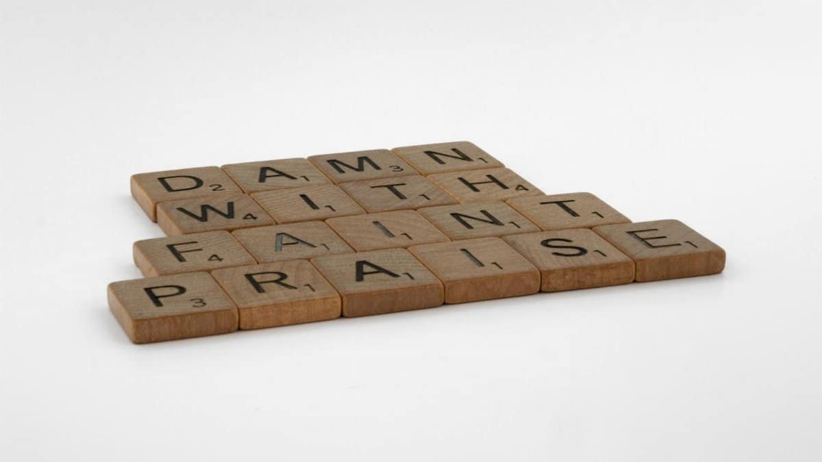 Close-up of wooden scrabble tiles spelling 'Damn with faint praise' on white surface.