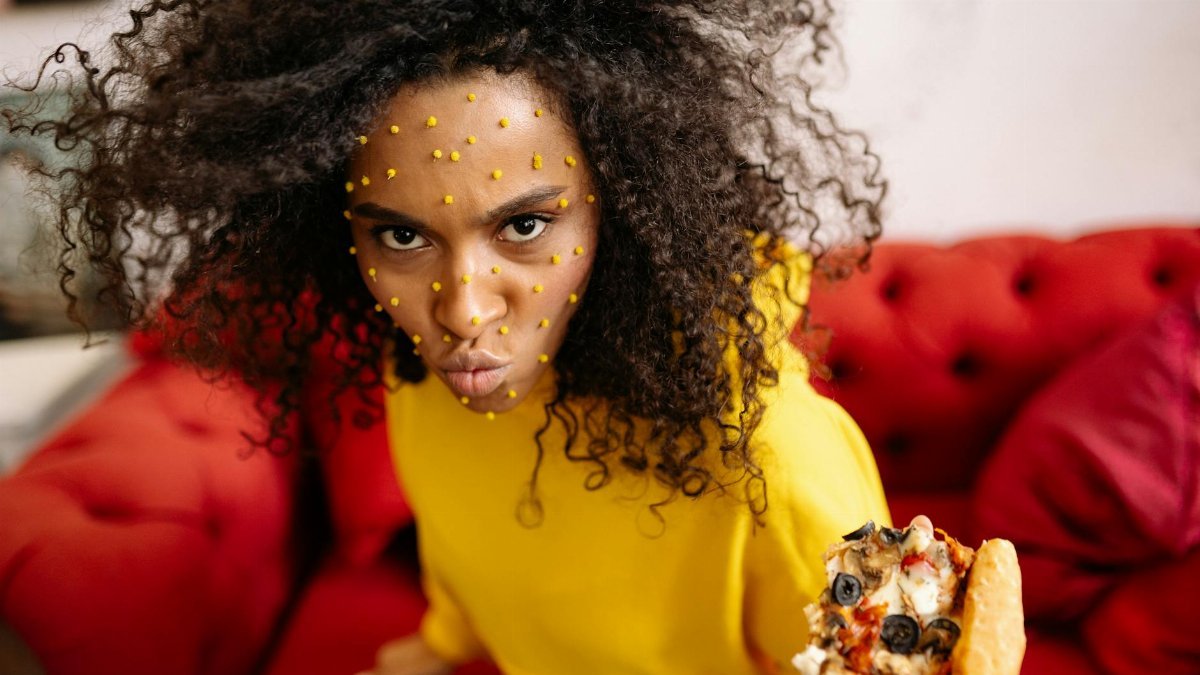 Playful woman with curly hair and dots, holding pizza on a red couch.