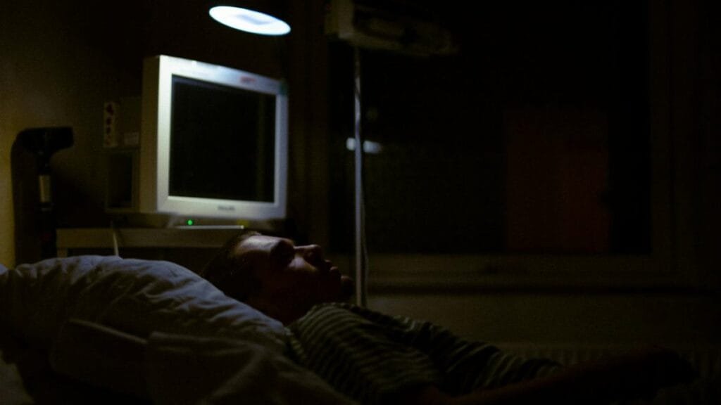 Man lying on a hospital bed under dim light, with medical equipment visible.