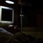 Man lying on a hospital bed under dim light, with medical equipment visible.