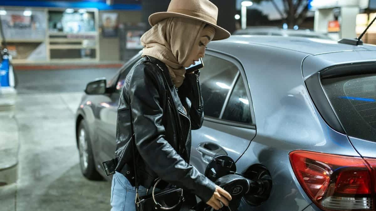 A woman in a hijab and hat refuels her car at a gas station, using a fuel pump.