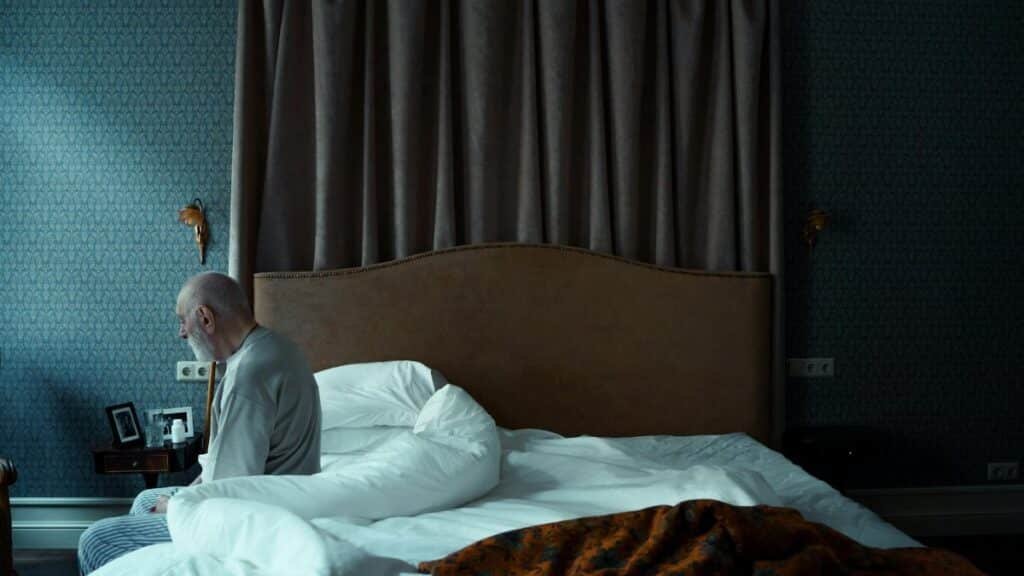 A senior man in pajamas sits on a bed, conveying solitude and reflection in a dark room.