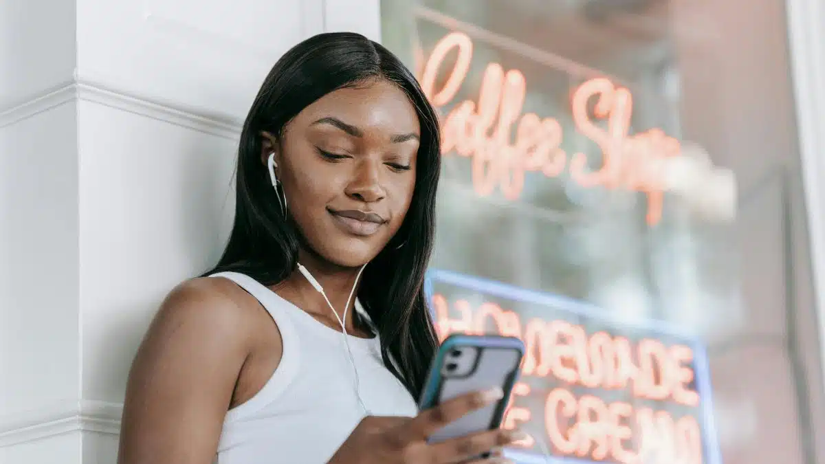 Smiling woman with earphones using smartphone outside a coffee shop.