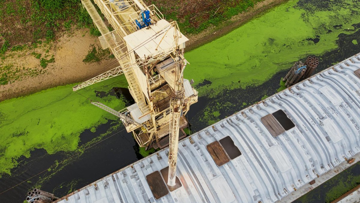 Aerial shot capturing an industrial crane over a river with green algae, highlighting industrial impact on nature.