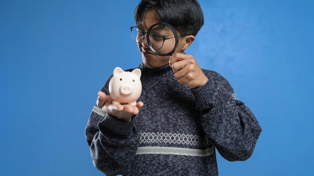 A person closely examines a piggy bank with a magnifying glass, symbolizing financial scrutiny.