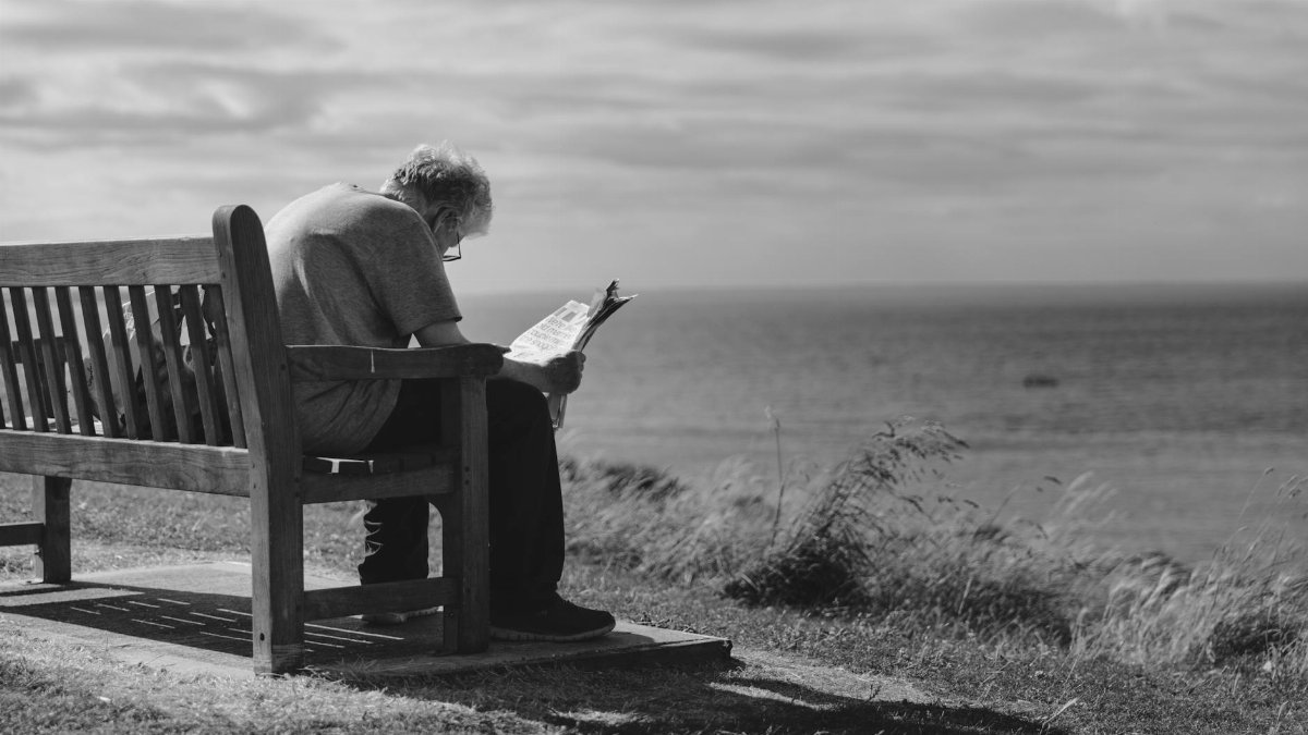 A contemplative scene of an elderly man reading a newspaper on a seaside bench with cloudy skies.
