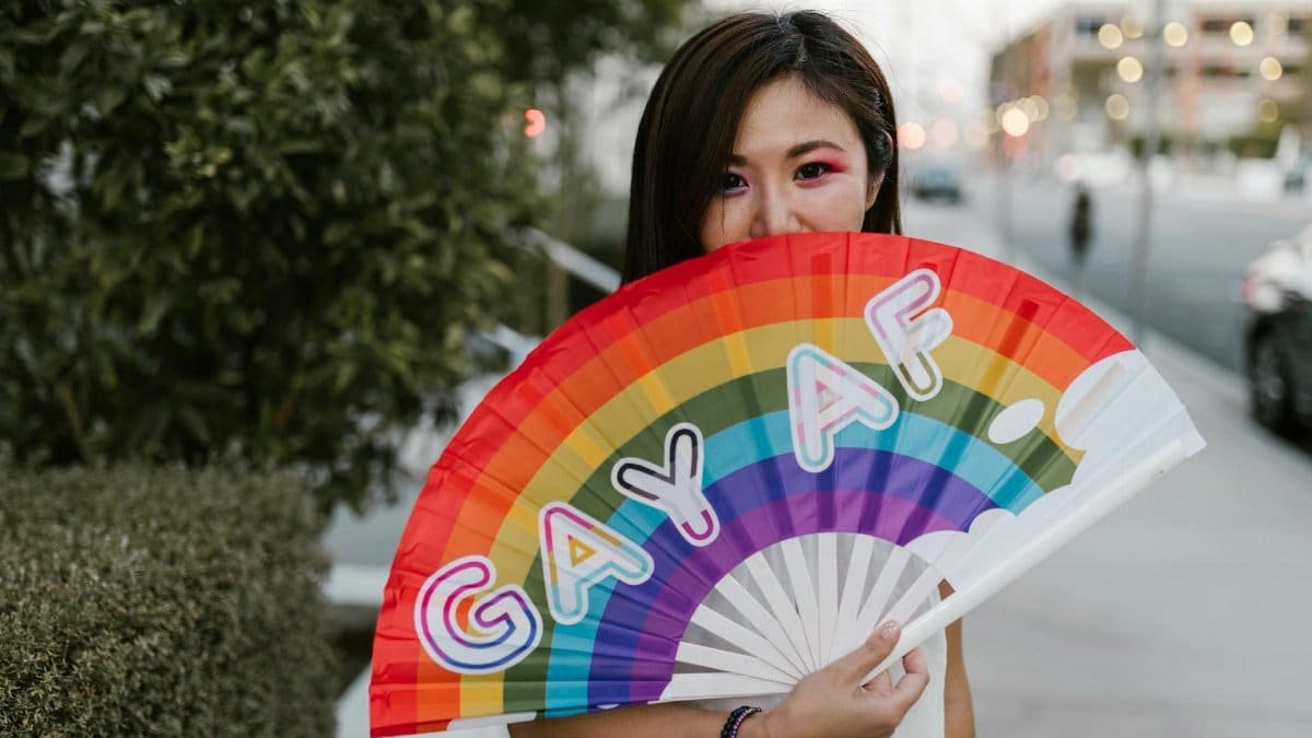 A colorful handheld fan with rainbow colors and the phrase 'GAY AF' held by a woman outdoors.