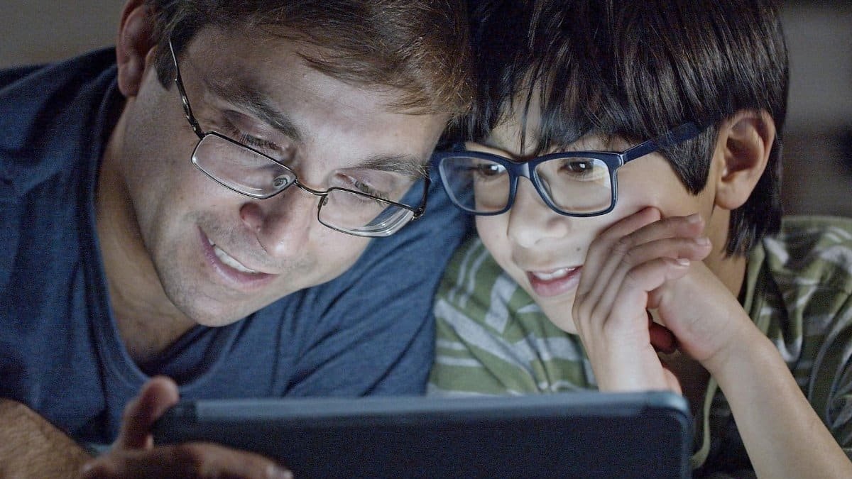 A father and son joyfully share a moment together, watching a tablet indoors.