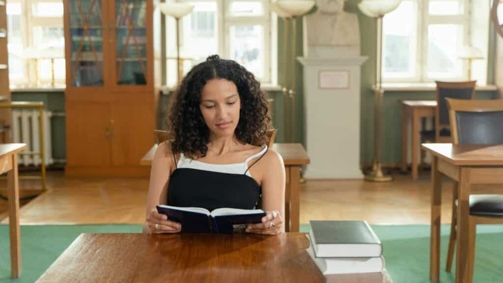 A young woman reads attentively in a well-lit library, surrounded by books.