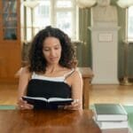A young woman reads attentively in a well-lit library, surrounded by books.