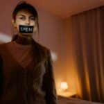 A woman stands in a dimly lit bedroom with tape reading 'Speak' over her mouth, symbolizing silence and oppression.