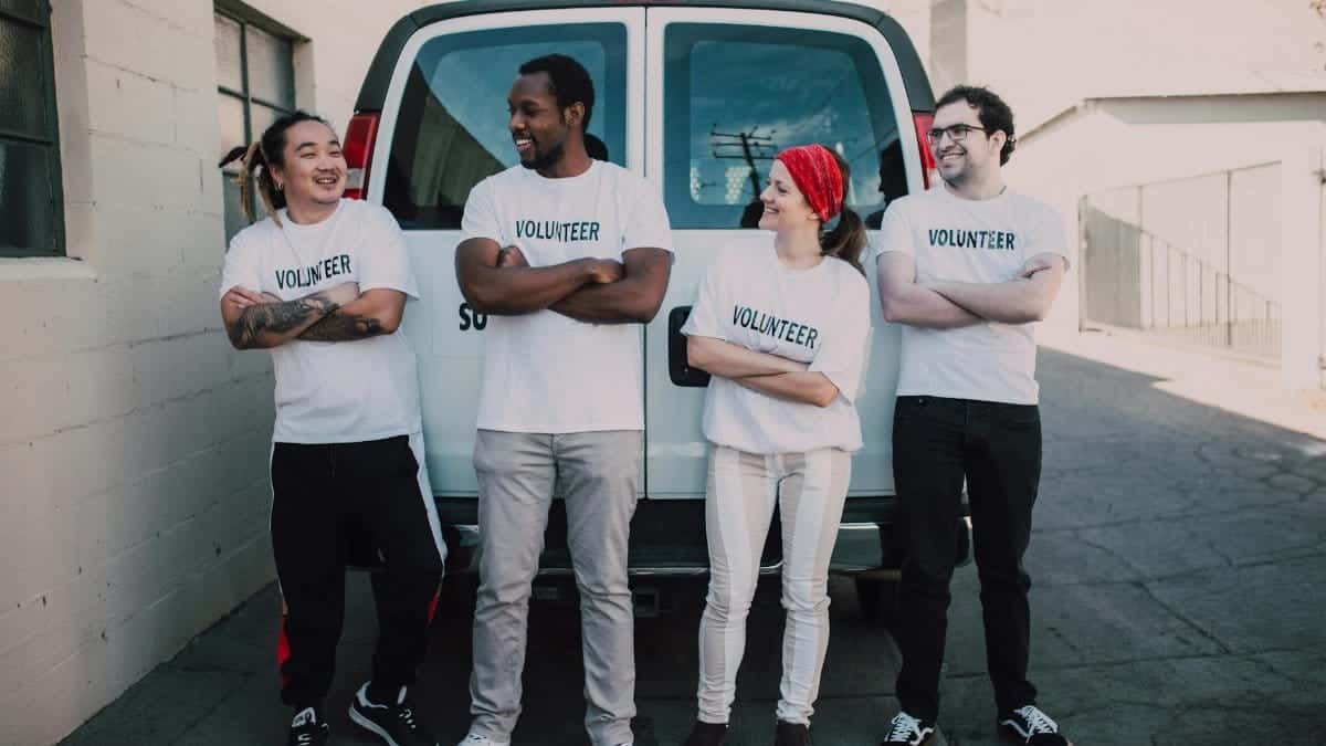 Four diverse volunteers in matching shirts smiling and standing by a van during a community service event.
