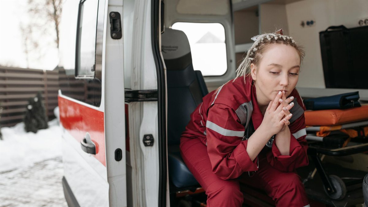 A female paramedic sits pensively beside an ambulance, reflecting on duty.