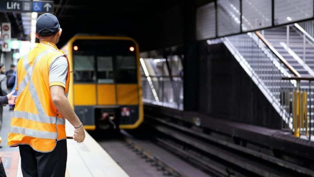 Railway staff in safety vest on platform as train arrives at Sydney station.