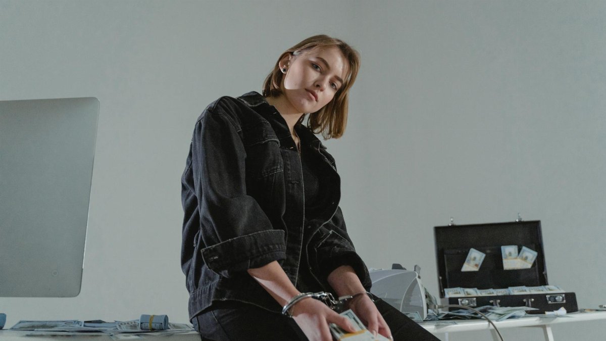 Young woman in black jacket handcuffed with cash, conceptual indoors setting.