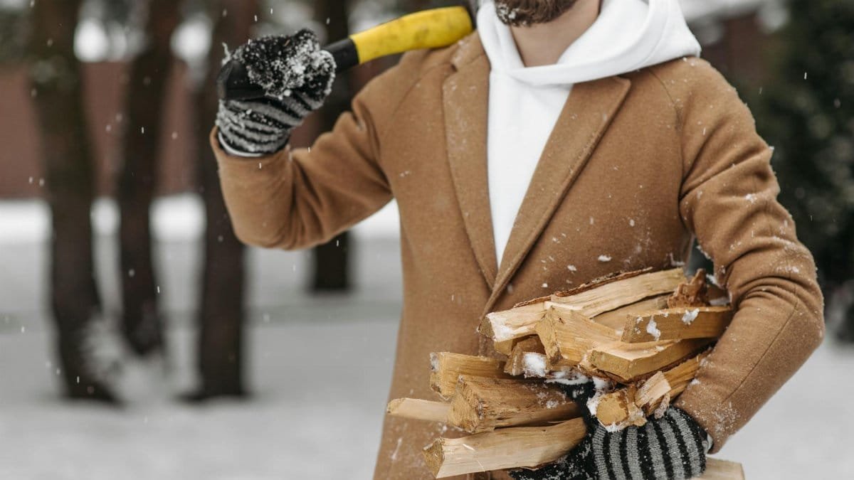 A man carries firewood in a snowy forest, wearing a coat and gloves during snowfall.