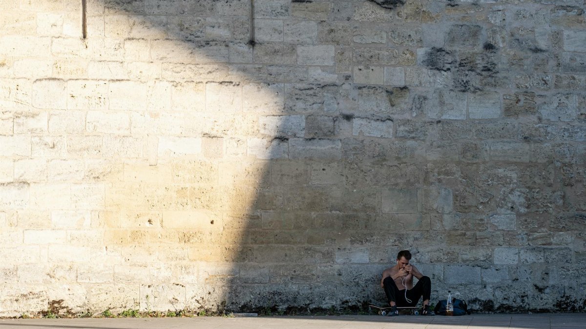 A young man sits with his skateboard against a sunlit stone wall, capturing a moment of solitude.