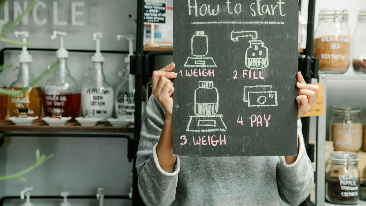 Person holding a chalkboard with shopping steps in a refill shop with glass containers on shelves.