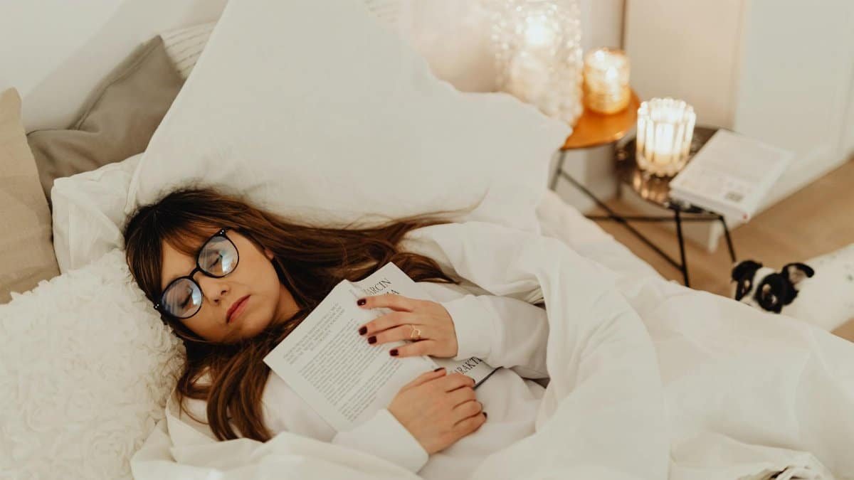 Woman with glasses asleep holding a book in a cozy bedroom setting with candles.
