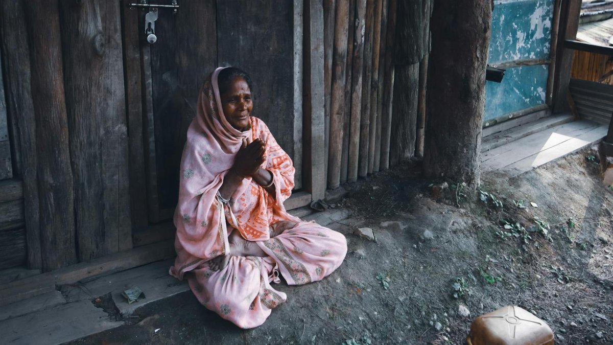 Elderly woman in a headscarf praying outside a rustic wooden building.