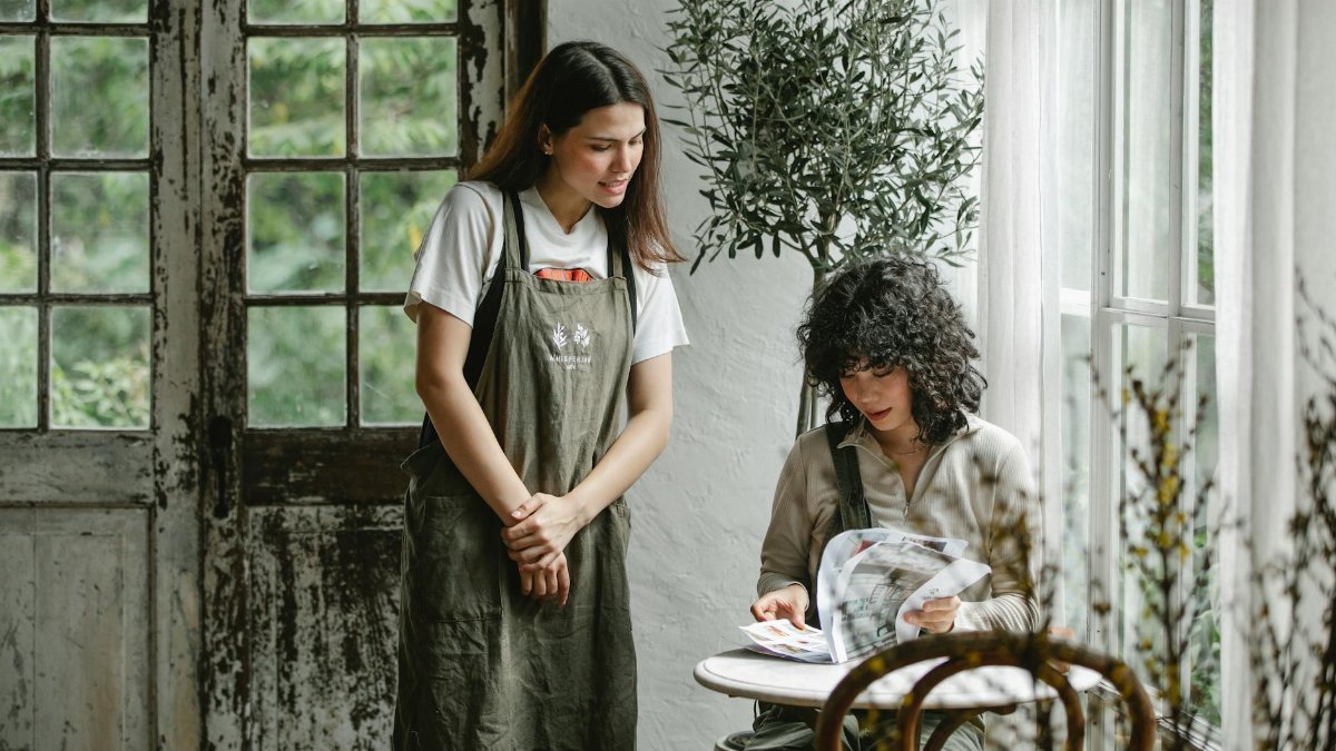 A waitress assists a customer reading a menu in a light-filled rustic cafe.