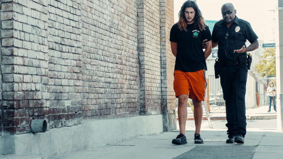 Police officer walking with handcuffed man along urban street with brick wall.