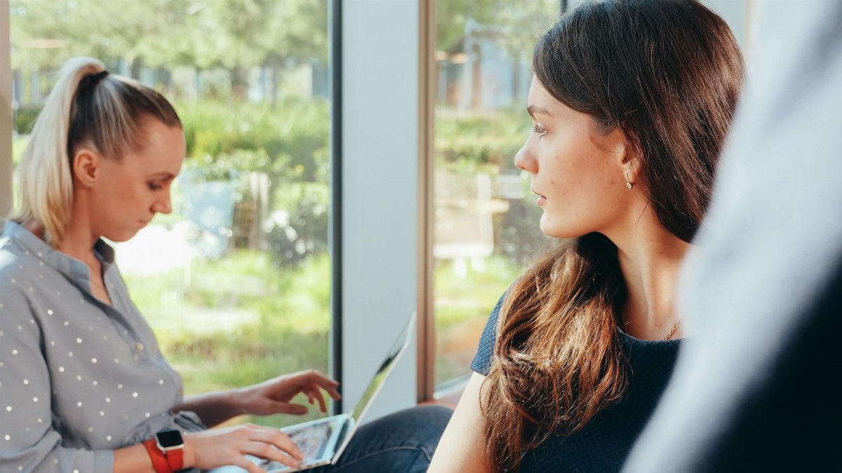 Two women sit by a window, one working on a laptop, the other looking outside.