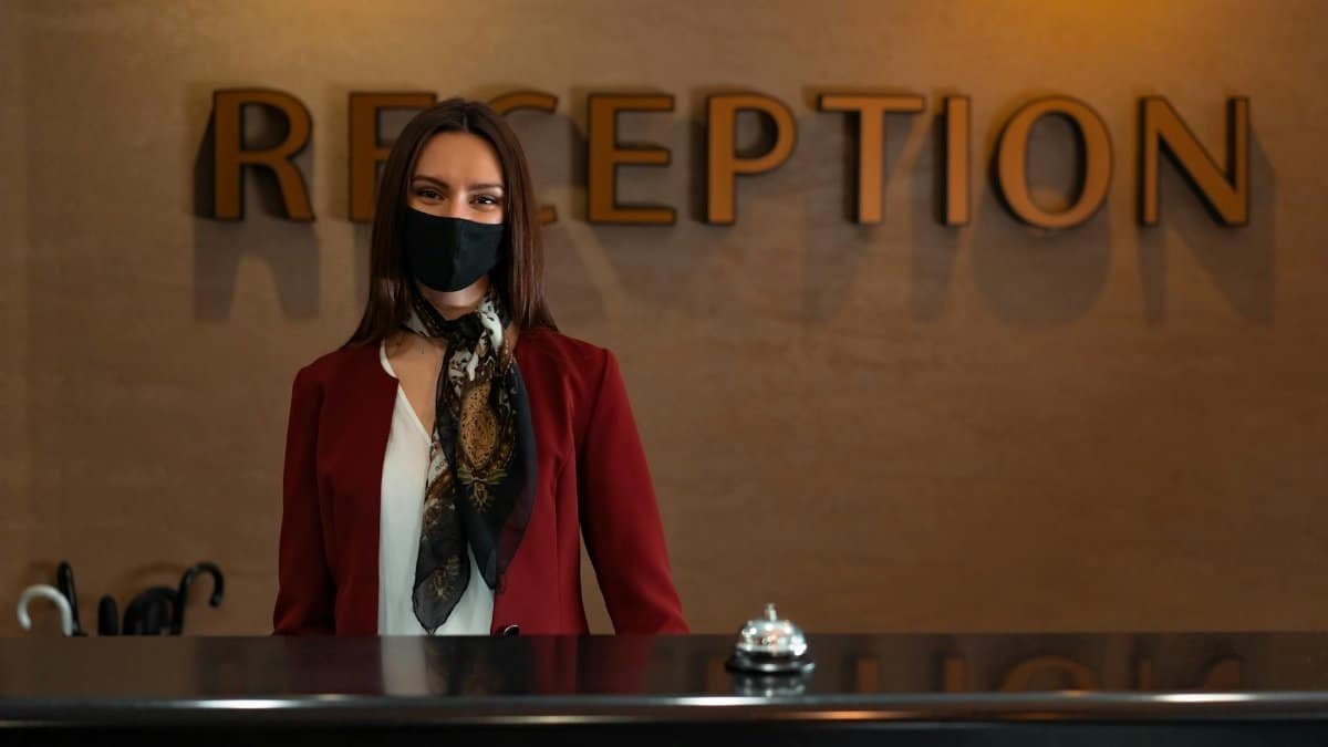 Woman in mask at hotel reception desk ready to assist guests.