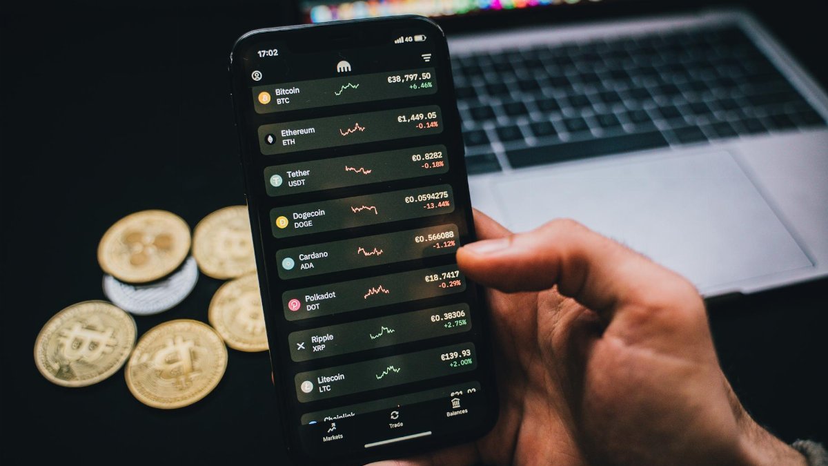 A person checks cryptocurrency values on a smartphone with a laptop and Bitcoins nearby.
