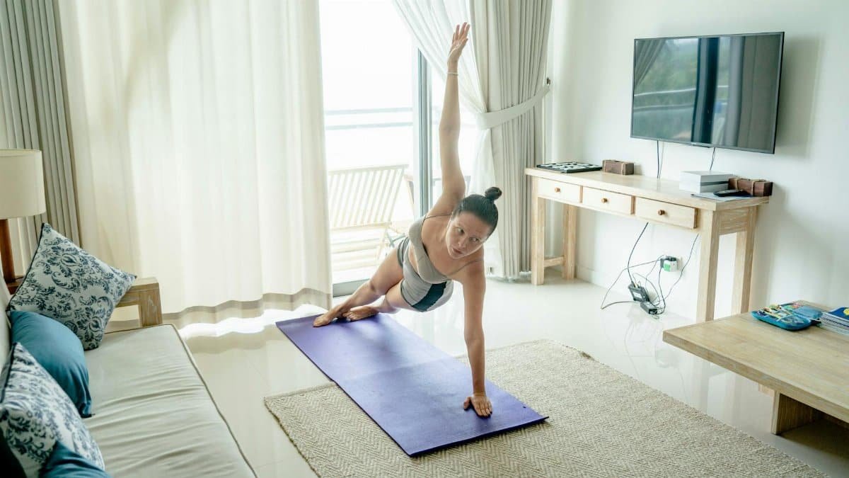 A woman practices yoga in a bright living room, performing a side plank on a mat.