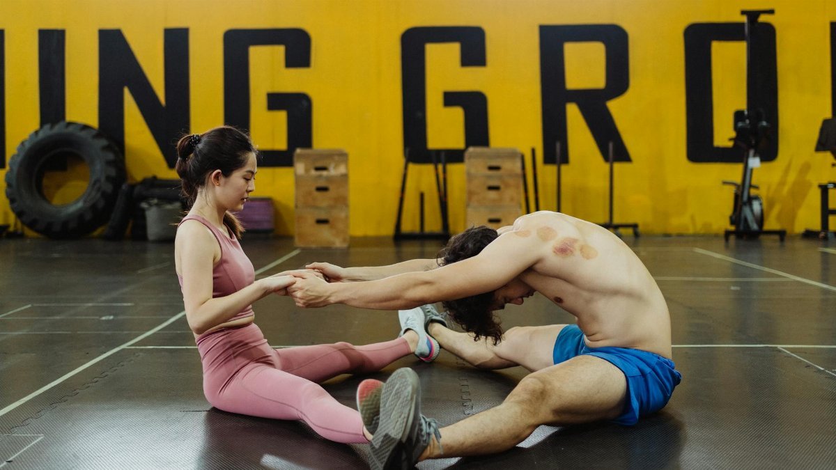A man and woman performing partner stretching exercises in an indoor gym setting.