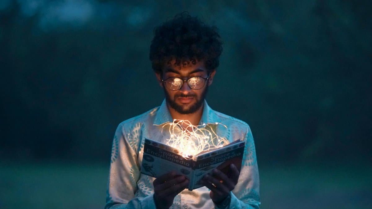 A man with glasses reads an illuminated book outdoors during twilight.