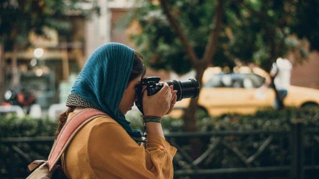 Female photographer with camera capturing street scenes, wearing casual attire and headscarf.